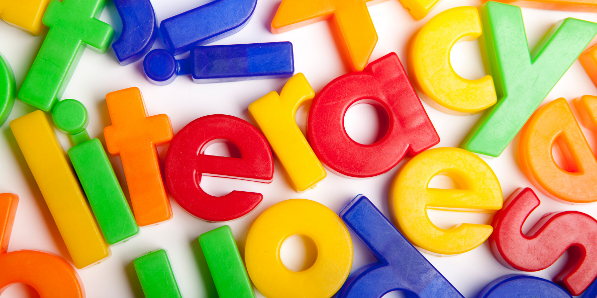 Colorful plastic letters scattered on a white surface, with the word 'literacy' formed in the center using red, yellow, green, blue, and orange letters.
